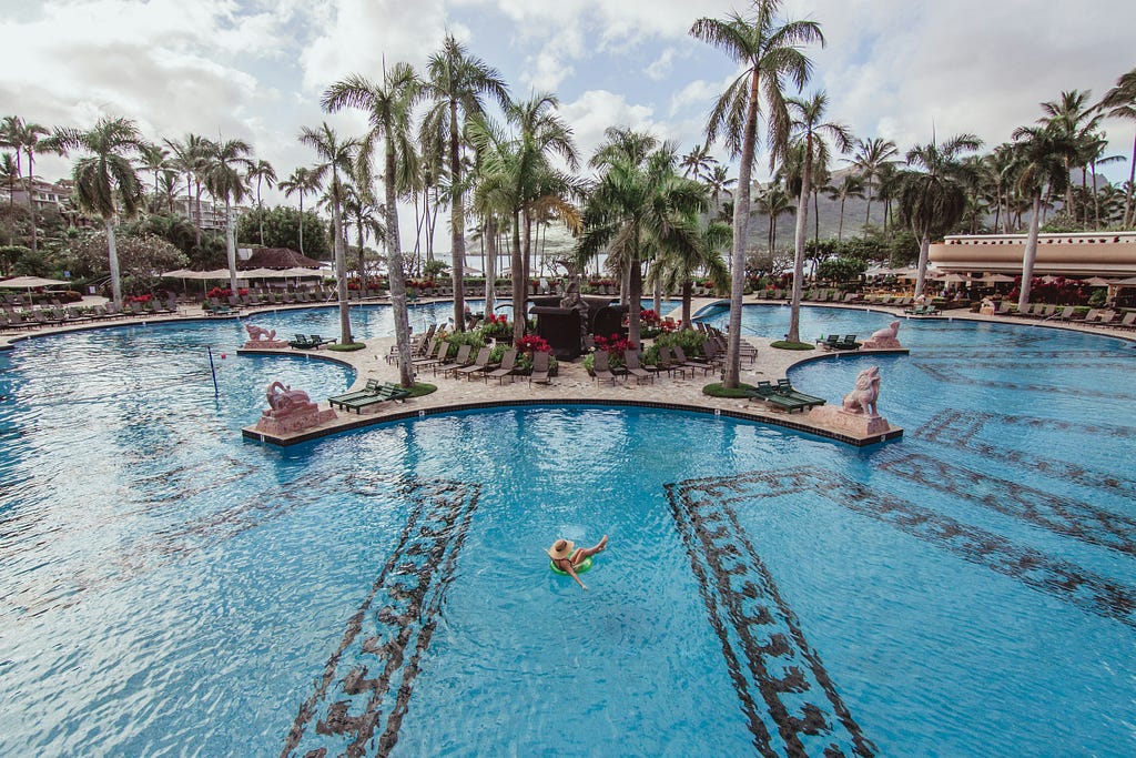 A holiday maker floats in a large swimming pool. The pool has a central island with palm trees on it.