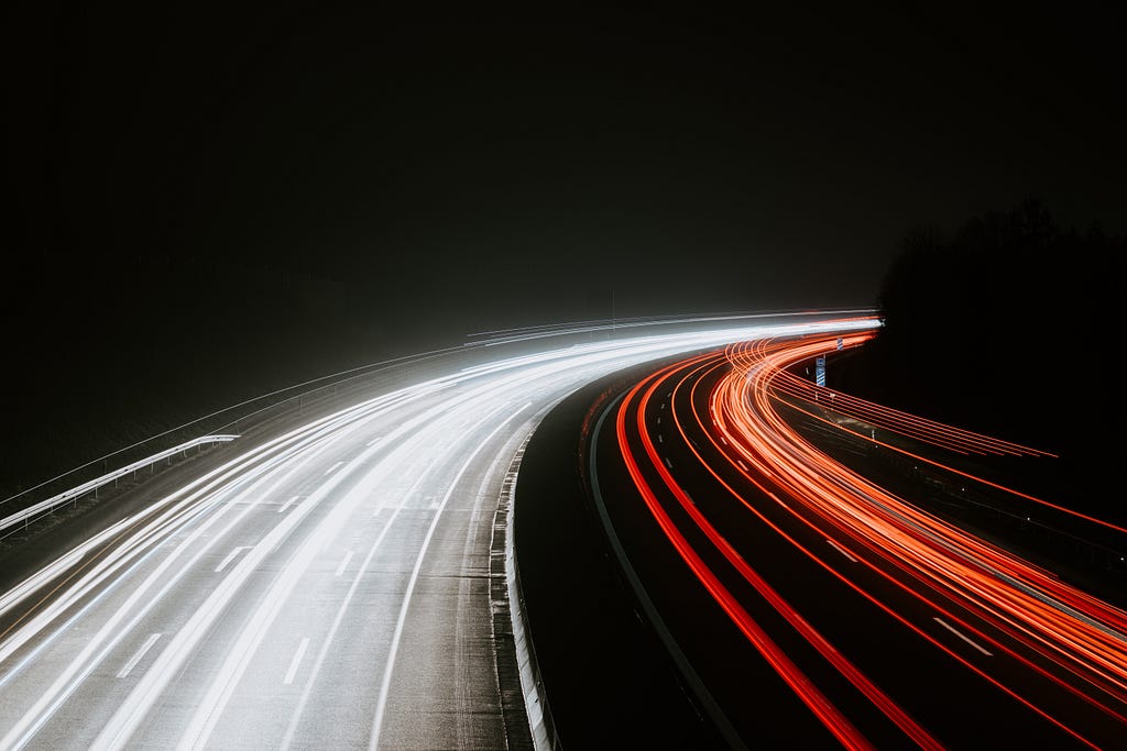 A shot of a highway at night with a streak of white lights on the left hand side of a highway and a streak of red lights on the right hand side of a highway.