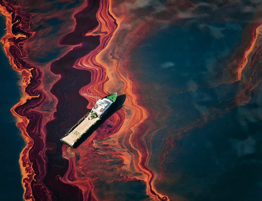 An arial photo of the BP oil spill, long streaks of orange and red oil plumes stretch across the ocean with a tanker seen navigating throught it.