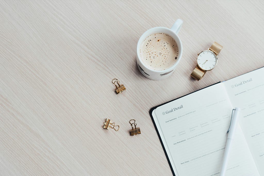 Caderno de anotações com uma caneta encima. Ao lado, um café, um relógio e alguns acessórios de papelaria metálicos.