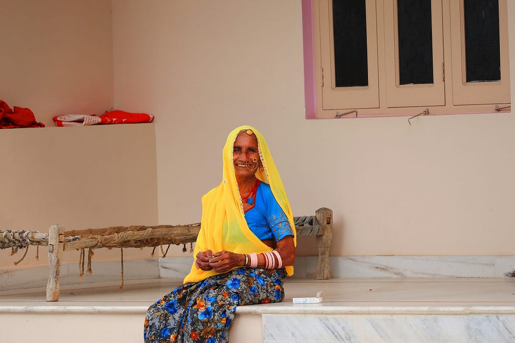 An elderly Indian woman wearing a bright yellow veil and blue blouse, sitting on a porch with a warm smile, traditional bangles on her arms, and a charpai (woven cot) beside her.