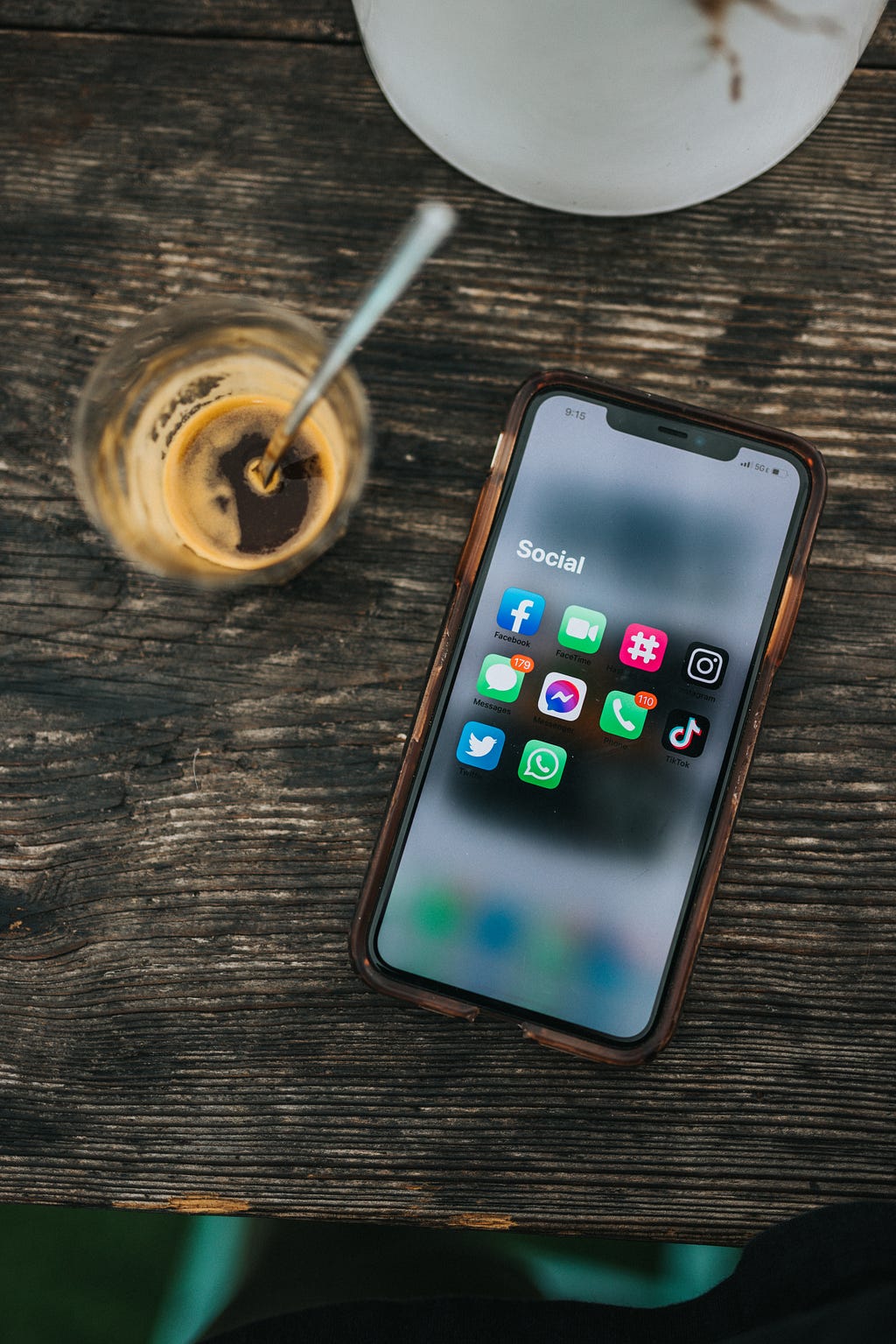An iPhone laying flat on a table, showing a homescreen view of a few different social apps (including Facebook, Twitter, TikTok, Instagram). The phone is lying next to an almost-empty glass of espresso.