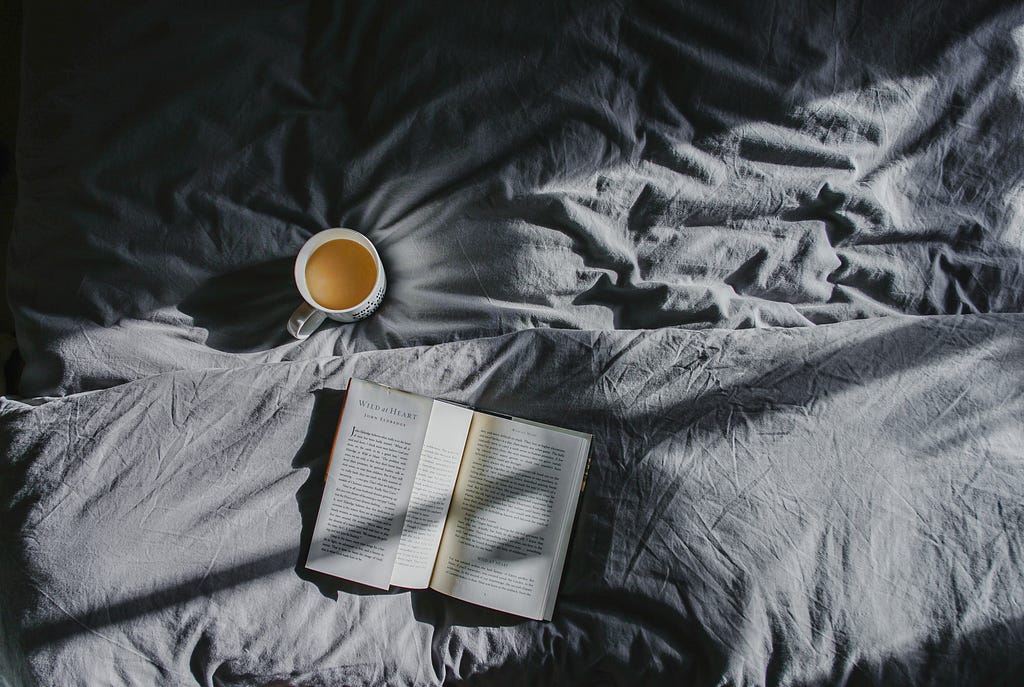 A bird’s eye view of a bed with gray bedding. On the bed is a cup of coffee in a white mug, and close by is an open book.