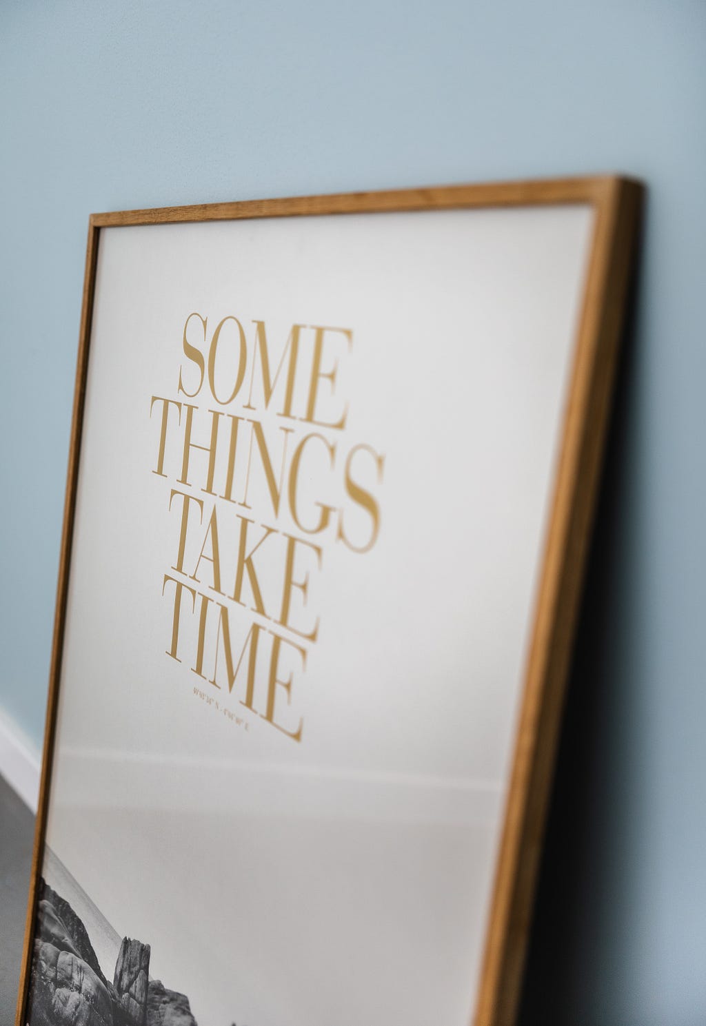 Quadro de fundo branco com moldura amadeirada com a frase “Some things take time” em dourado e letras em caixa alta