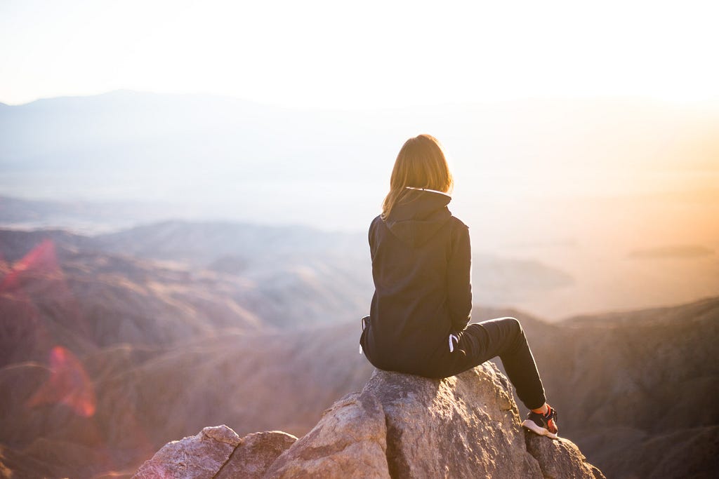 A woman in a black jacket and jeans sits at the top of a mountain, looking into the sunset. You can see the back of her head.