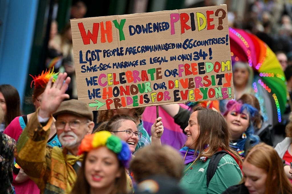 People of all ages and genders colourfully dressed and celebrating, in a parade. In the centre of the picture, people hold a banner reading: “Why do we need Pride? LGBT people are criminalised in 66 countries! Same sex marriage is legal in only 34! We celebrate our freedom. We honour our diversity. Pride is for everyone!”