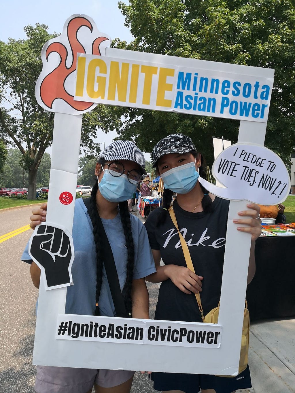 Two young people wearing face masks and baseball caps holding up a large cardboard frame with the words “IGNITE Minnesota Asian Power” and “I pledge to vote Tuesday, November 2!”