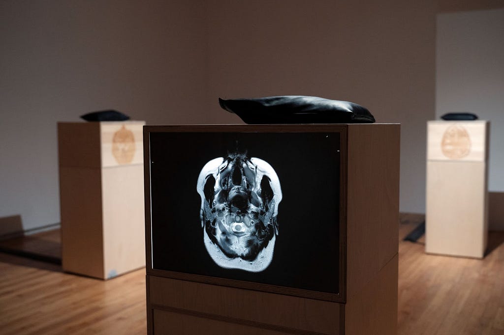 Three plinths are in view in a white-wall art gallery, spaced apart with one closest up showing the most detail. On each plinth is a vertically placed image; each shows a different brain pattern in the top-down view of a MRI scan. The closest image has the brain scan details in white on a black background, and within the contours of the brain shape is overlaid a portrait photo of a brown woman facing front with head tilted back and eyes closed. In ritual adornment, she presents a spiritual pose.