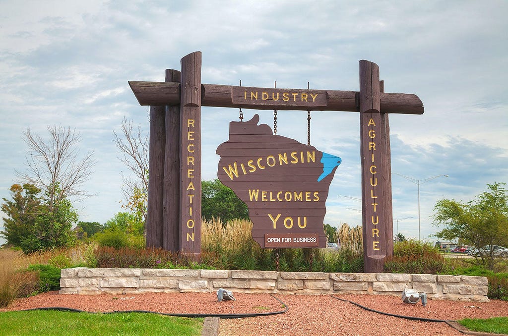 Wooden welcome sign at a state border featuring the outline of Wisconsin in dark brown against a lighter brown backdrop. Above and below the state outline are the words ‘INDUSTRY,’ ‘RECREATION,’ and ‘AGRICULTURE’ in gold letters, indicating key sectors. The center of the sign has ‘WISCONSIN WELCOMES YOU’ prominently displayed with a smaller ‘OPEN FOR BUSINESS’ caption below. The sign is flanked by posts with a rustic appearance, set in a well-maintained garden bed edged with stone under sky.