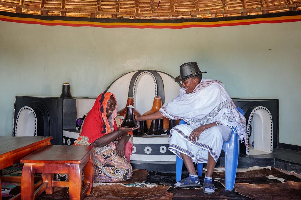 A tourist visiting the traditional Bahima home interacts with a woman holding a traditional container while seated on the floor.