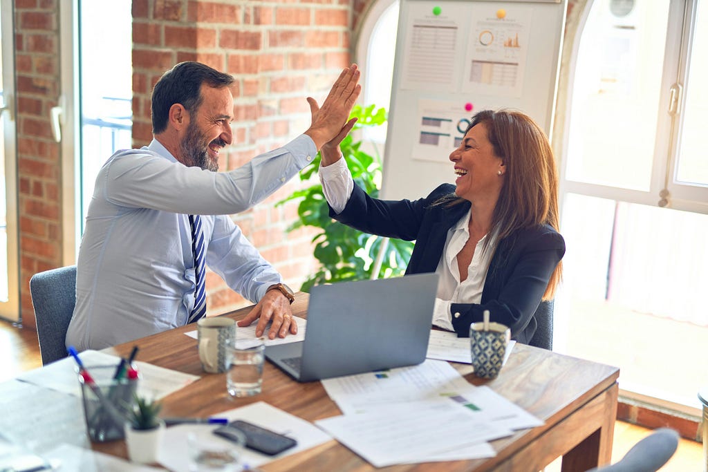 Two business people high-fiving over a desk which holds a laptop, papers, coffee mugs and other business paraphernalia. In the background there are windows letting in light and a whiteboard with graphs and data pinned to it