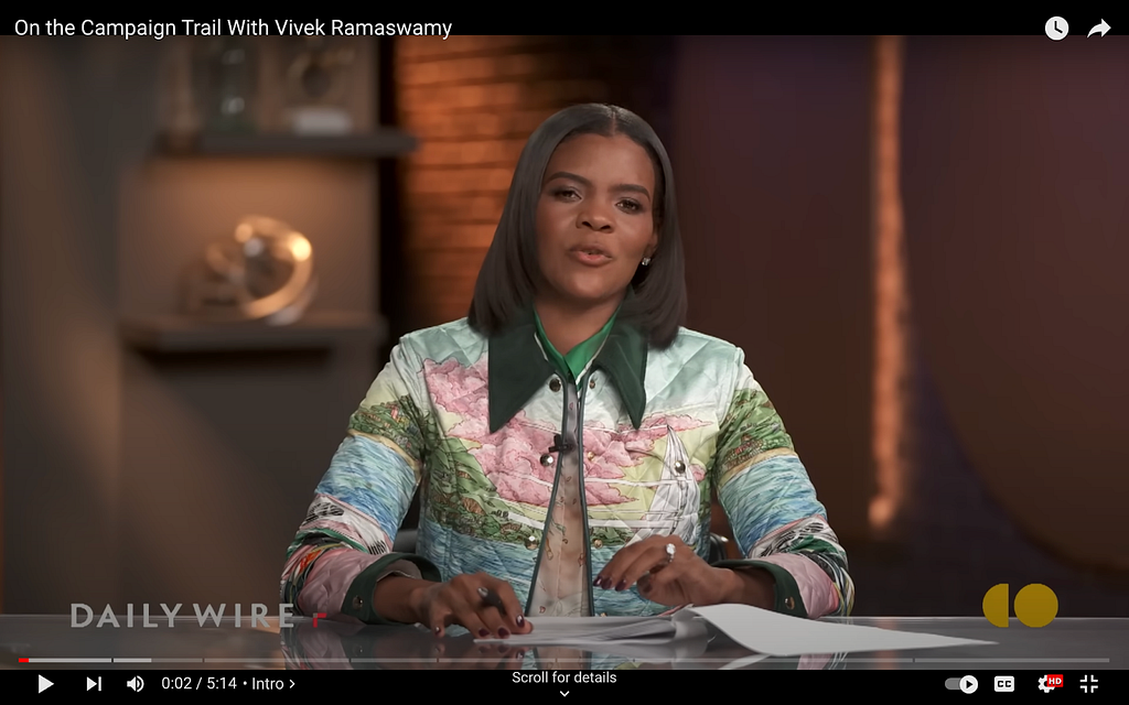 A screenshot from Candace Owens’s show on The Daily Wire. She’s in a quilted jacket with a forest green collar. There’s a scene printed across the jacket: In the background, green mountains and low, pink clouds. In the foreground, a sailboat gliding on a blue lake.