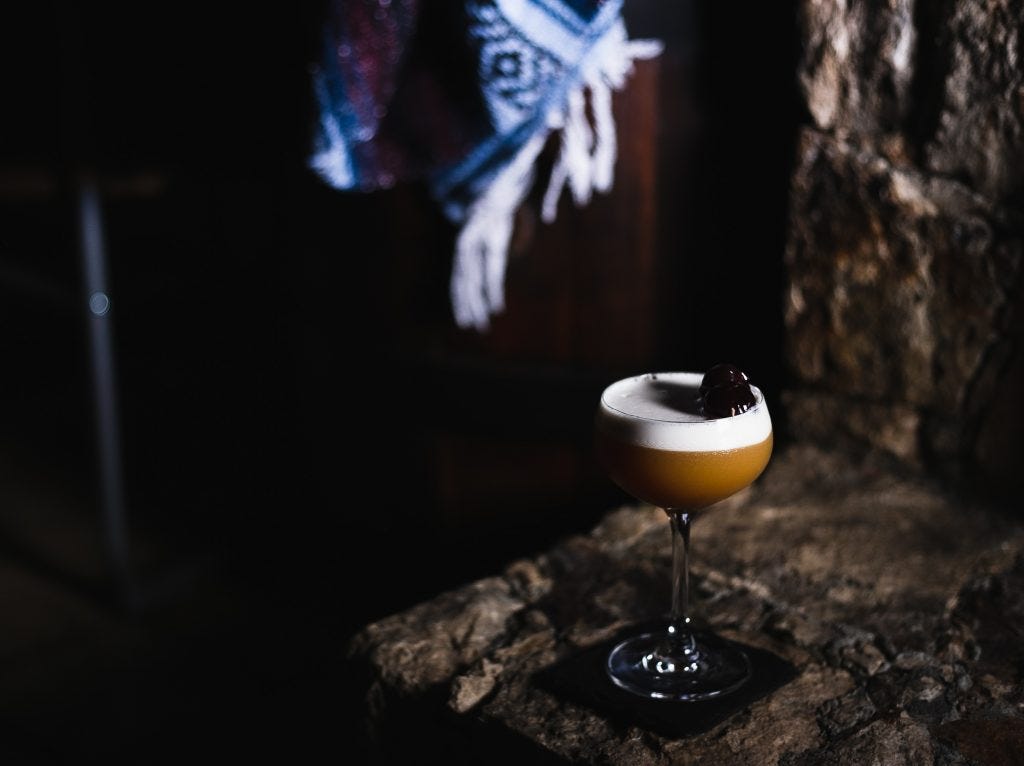 A long shot of an amaretto sour, with two cherries resting on the rim. A blanket is draped over a barrel in the background