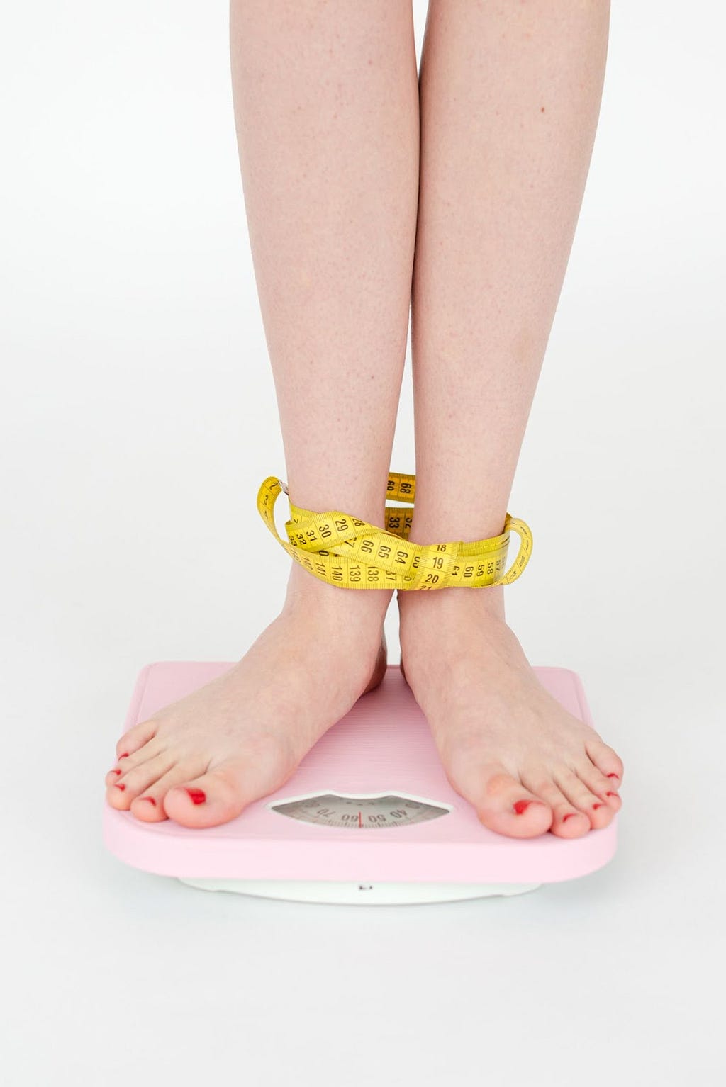 Image showing someone’s legs, standing on a weighing machine with an inch tape wrapped around their ankles.