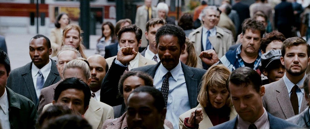 Cena final do filme À Procura da Felicidade, de 2006, em que o ator Will Smith está andando na rua, no meio de várias pessoas que usam roupas de trabalho, como ternos, roupas sociais e que tem no rosto um semblante desanimado. Ele, Will, destoa da cena ao levantar os braços comemorando.