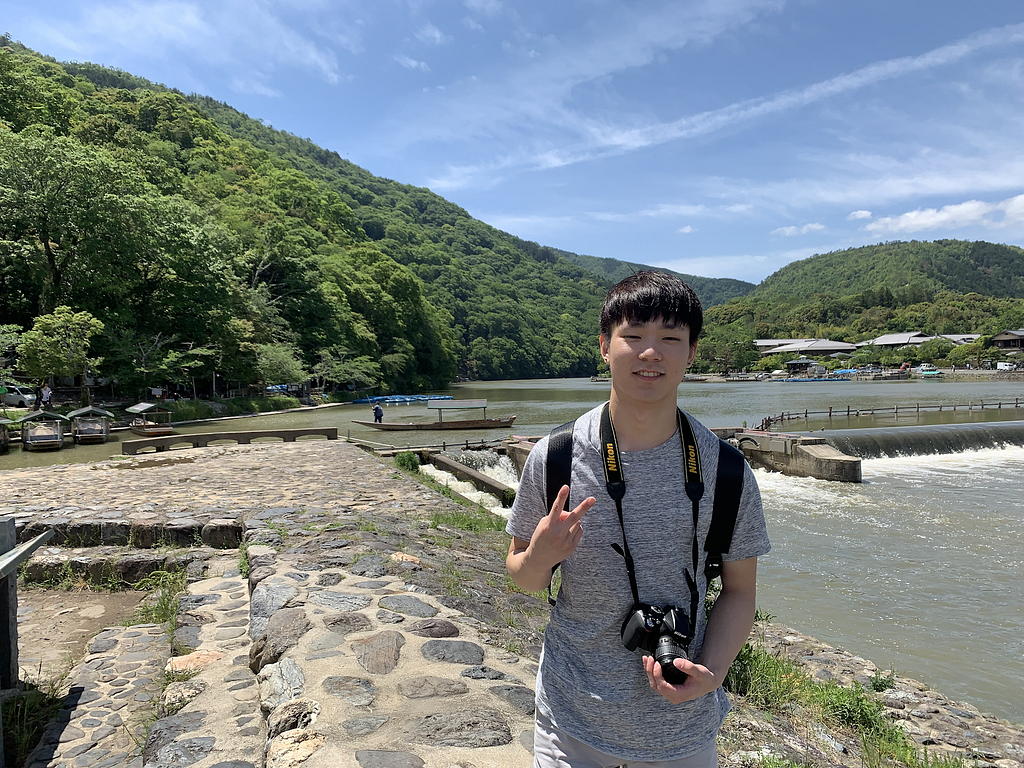 A person stands in front of a river with a small dam. He makes the peace sign with one hand, and holds a camera with other