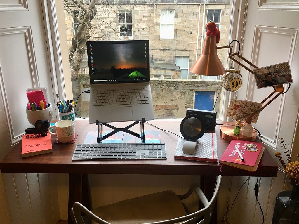A picture of Núria’s desk in a bay window, showing a laptop on a stand, external keyboard and mouse, speakers and task lamp.