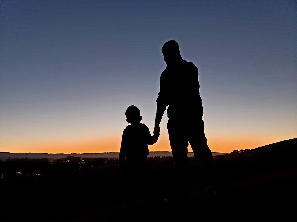 Silhouettes of a boy and his father, facing the city at sunset.