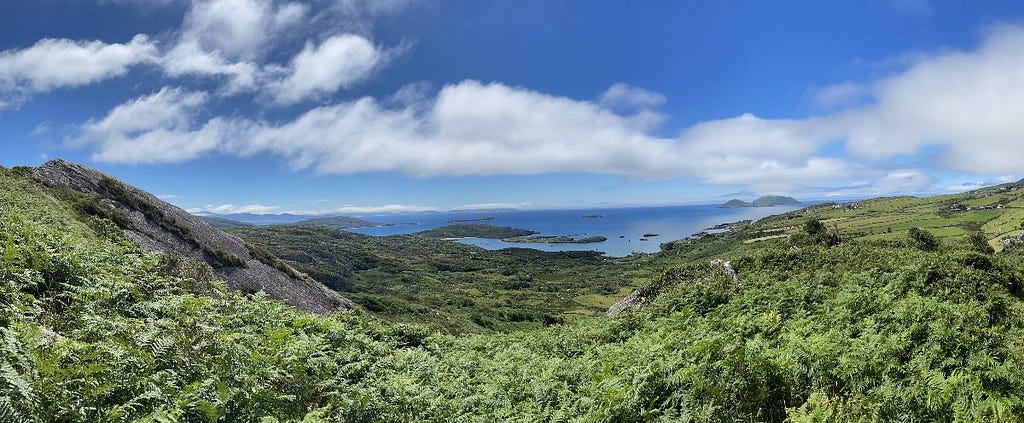 Photo from a hilltop in Derrynane, County Kerry, Ireland, with lush greenery in the foreground and small islands dotting the ocean in the distance. The sky is blue with puffy clouds.