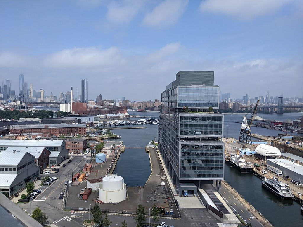 A photograph of a waterfront manufacturing area scattered with boats, industrial equipment, low-rise warehouses, and a skyline view.