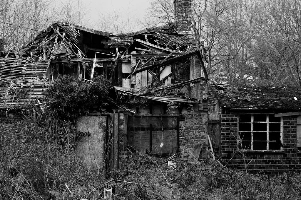 A black and white picture of an old, decaying home or barn. Rafters are exposed and toppling over. The building is barely still standing.