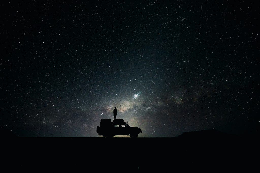 A man in desert standing on a Jeep looking at a starry night