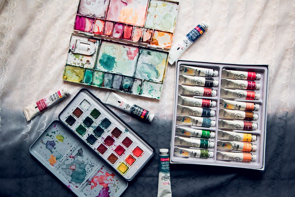 A top view photograph of a painting palette, a box of paint tubes and a tin box of watercolour pans