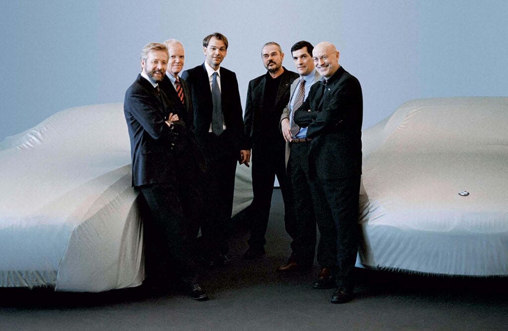 Ian Cameron con su equipo de BMW de finales de los 90, posando frente a dos coches cubiertos con lonas protectoras. Los individuos están vestidos con trajes formales, sugiriendo un entorno profesional.