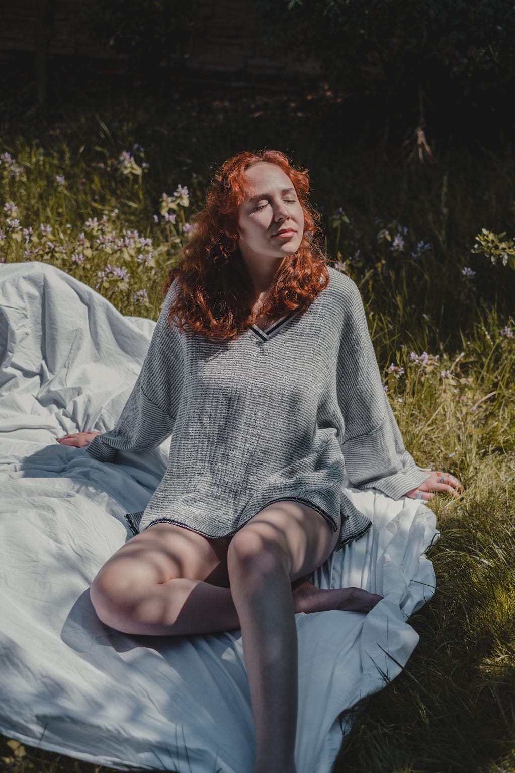 a female redhead soaking þe sun while sitting on a blanket in a meadow wearing grey.