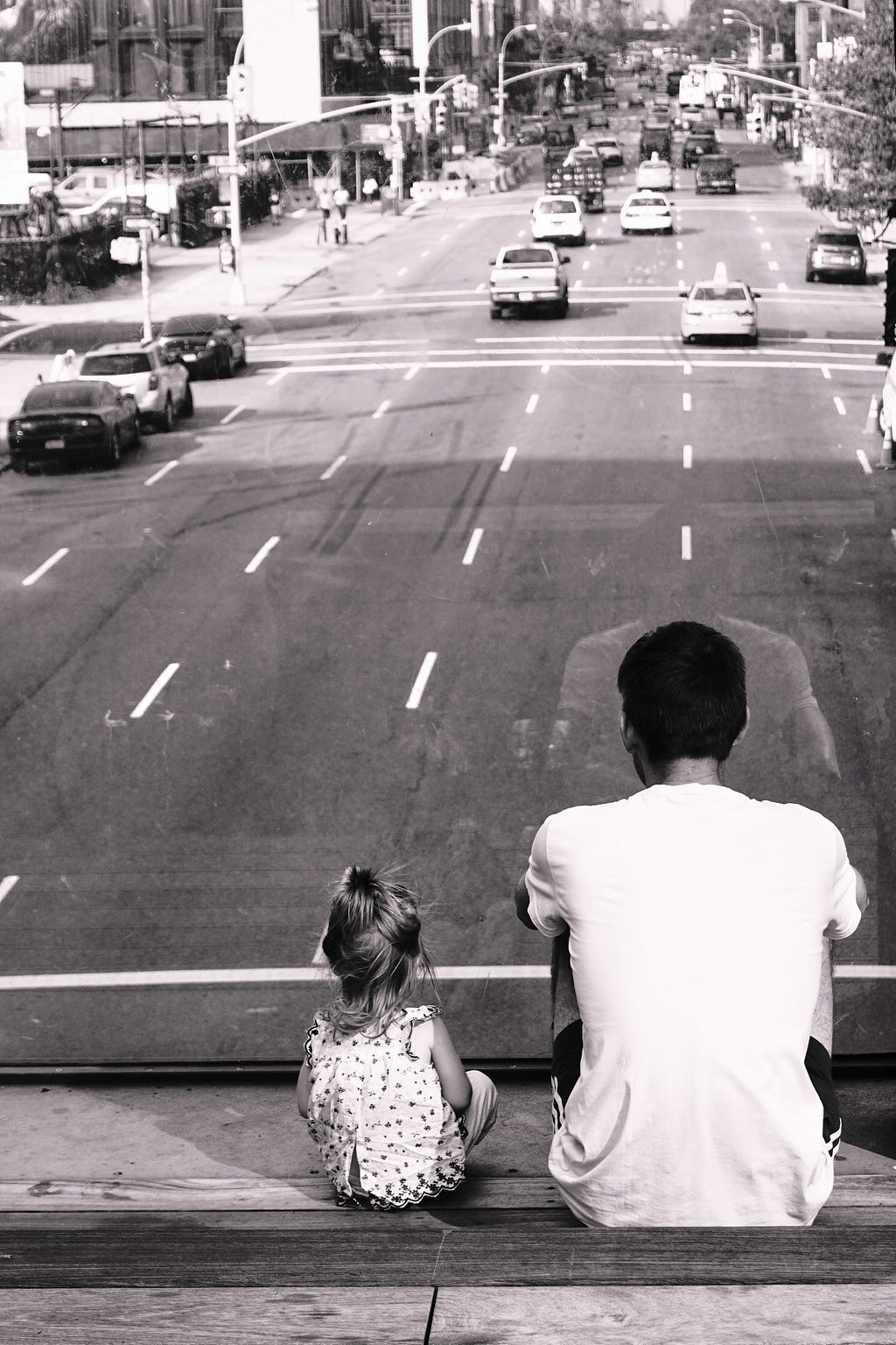 A black and white photo of a multi-lane city street in the background with the backs of a dad and little girl sitting above looking down at the road and distant traffic.