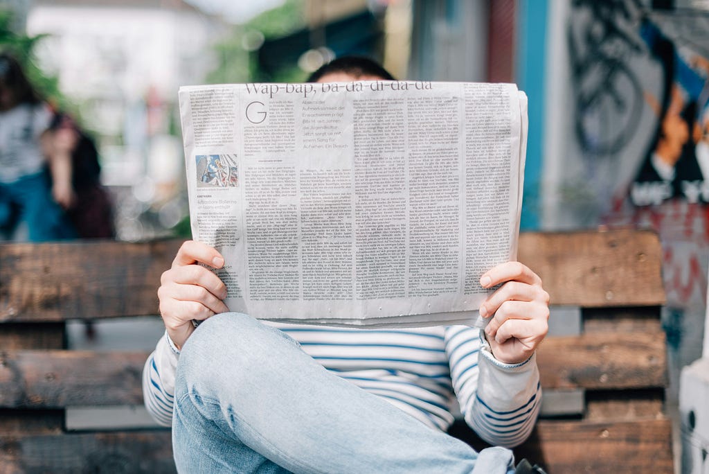 A reader holds up a newspaper to read on a park bench.