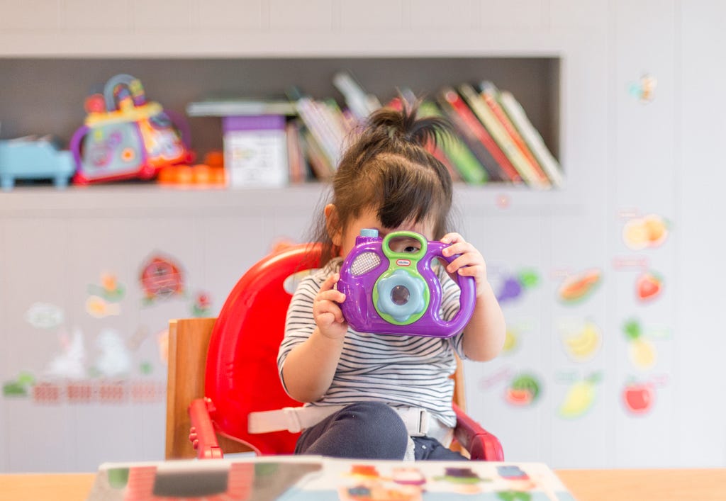 Set against the colorful backdrop of what may be a classroom wall with books, toys, and student work, a young child seated in a high chair with hair face obscured by a giant purple toy camera.