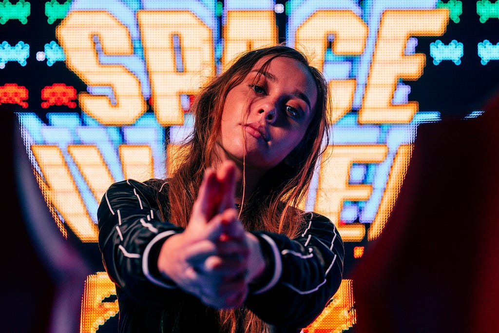 Photo on a young woman pointing directly at the camera with her hands in the form of a finger gun. Her background is a classic arcade screen.