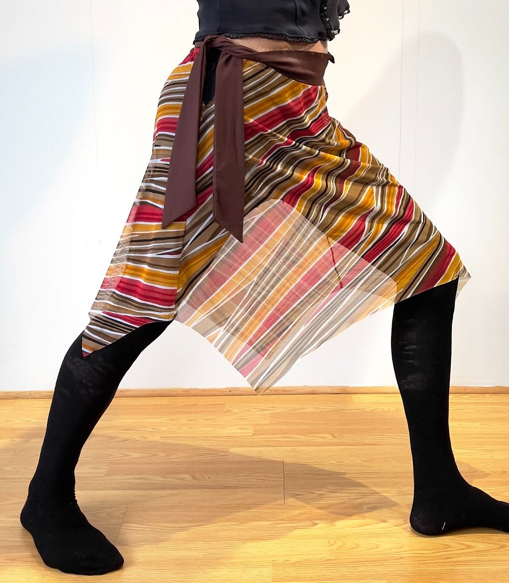 Me in a forward stance, with a red/orange/brown striped sarong, with brown ties at the top, and wearing black tights.
