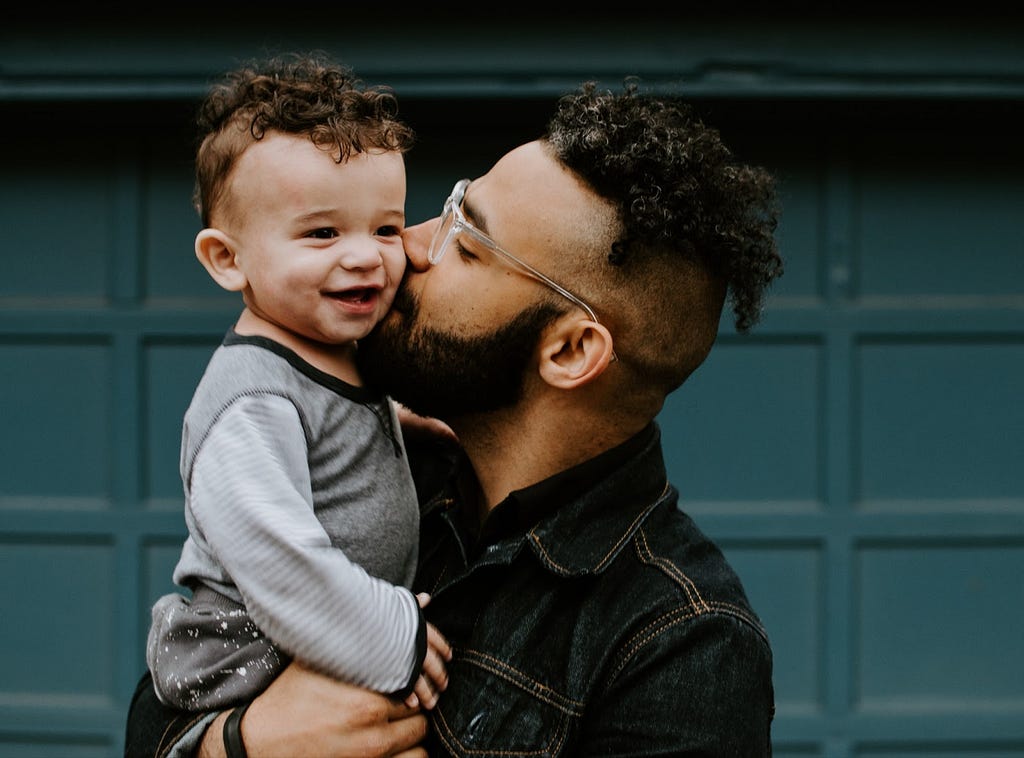 A young man with glasses and a beard holds his child on his shoulder and kisses him on the cheek. The child is smiling. They are standing in front of a green door.