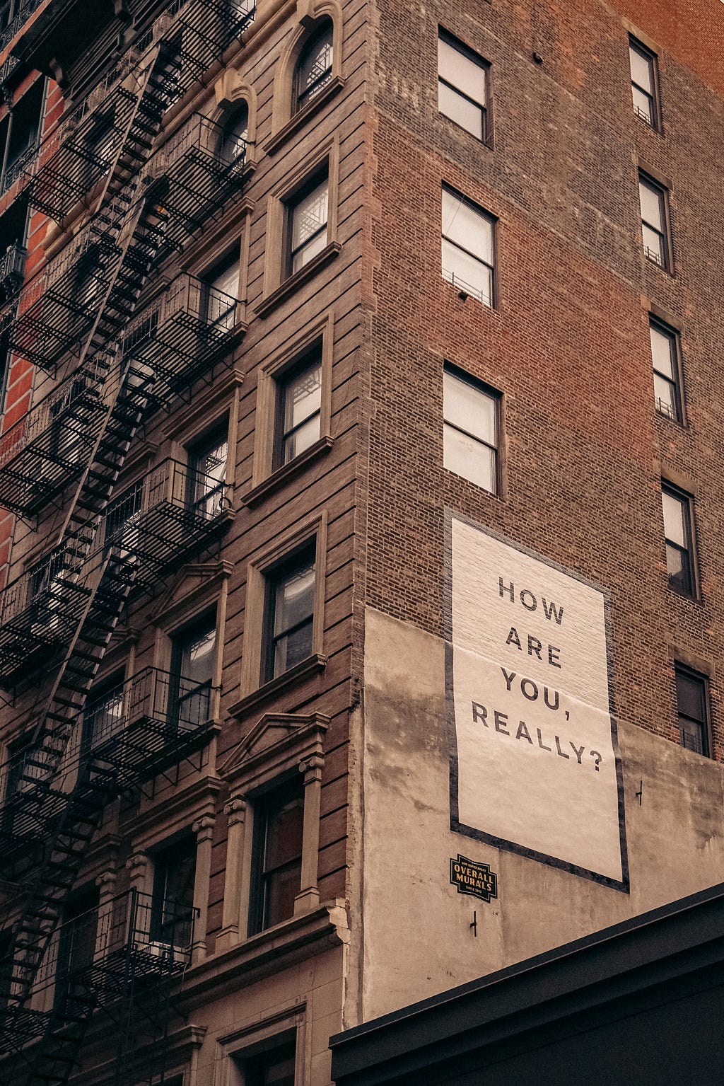 A brick apartment building with a mural on the side saying “How are you, really?”