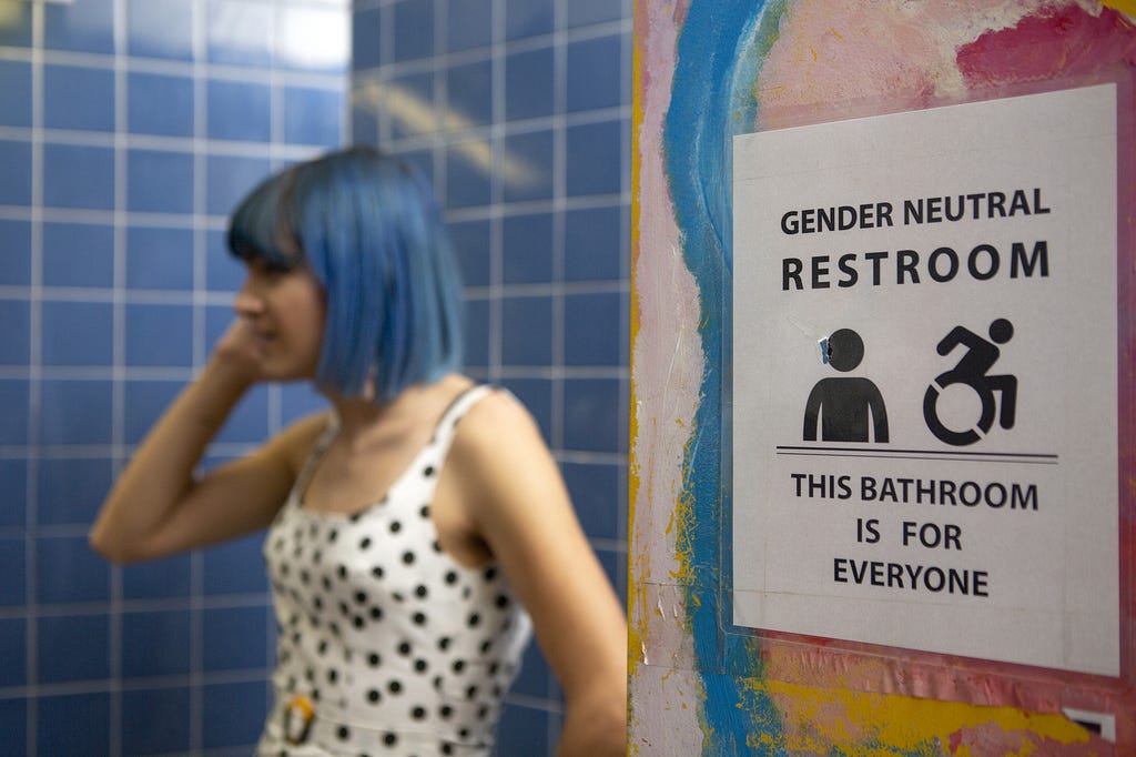 This picture of a white-passing non-binary person standing in a restroom next to a sign that reads “Gender Neutral Restroom.