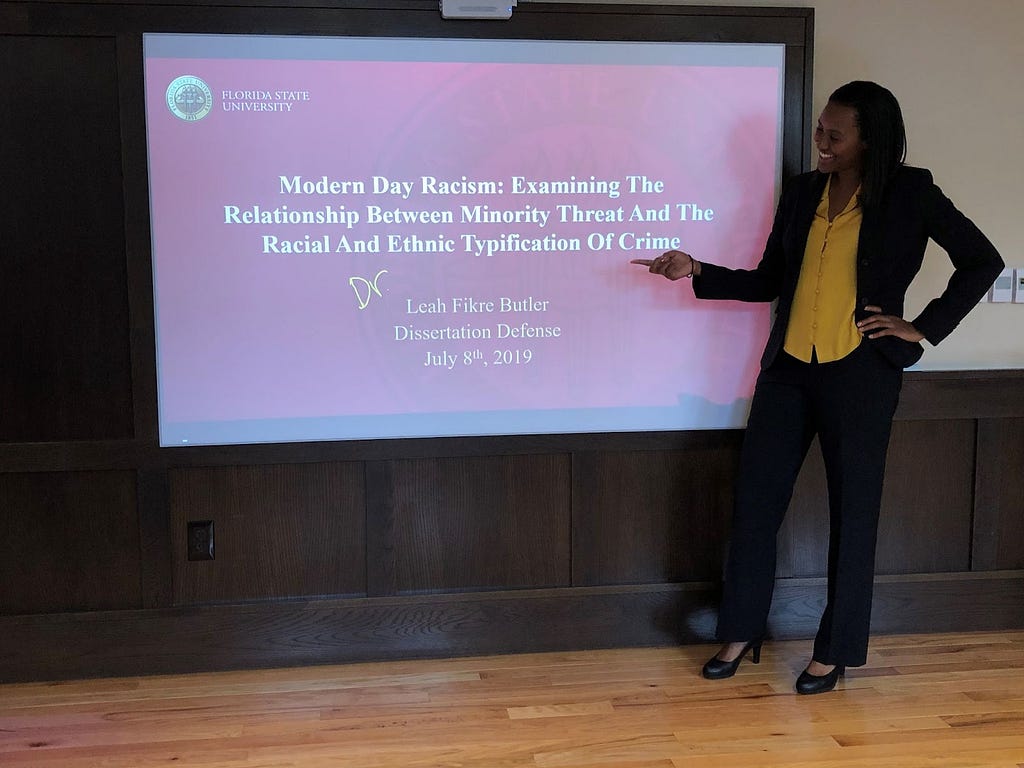 Leah Butler stands in front of a screen that shows her doctoral dissertation, “Modern Day Racism: Examining the Relationship Between Minority Threat and the Racial and Ethnic Typification of Crime.”