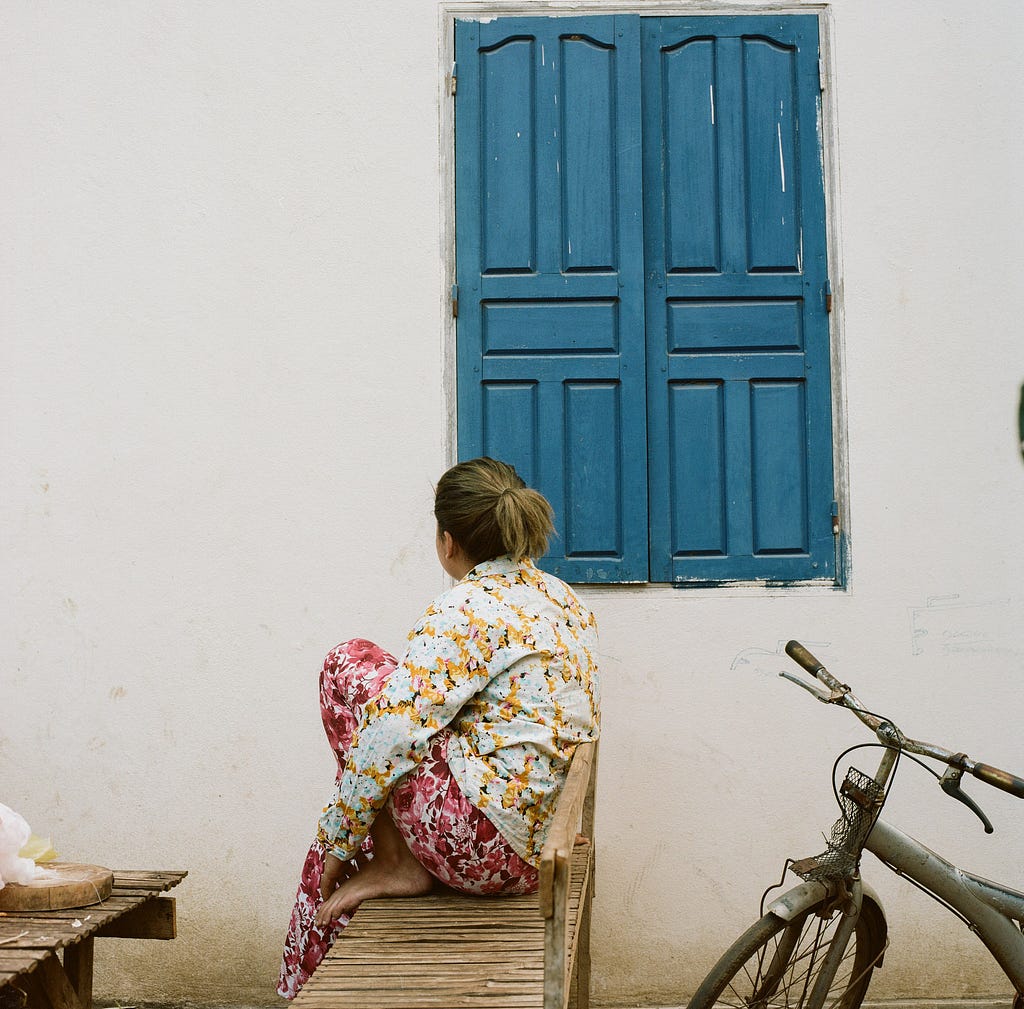 A female migrant worker sitting outside of her living quarters, facing the side near a window.
