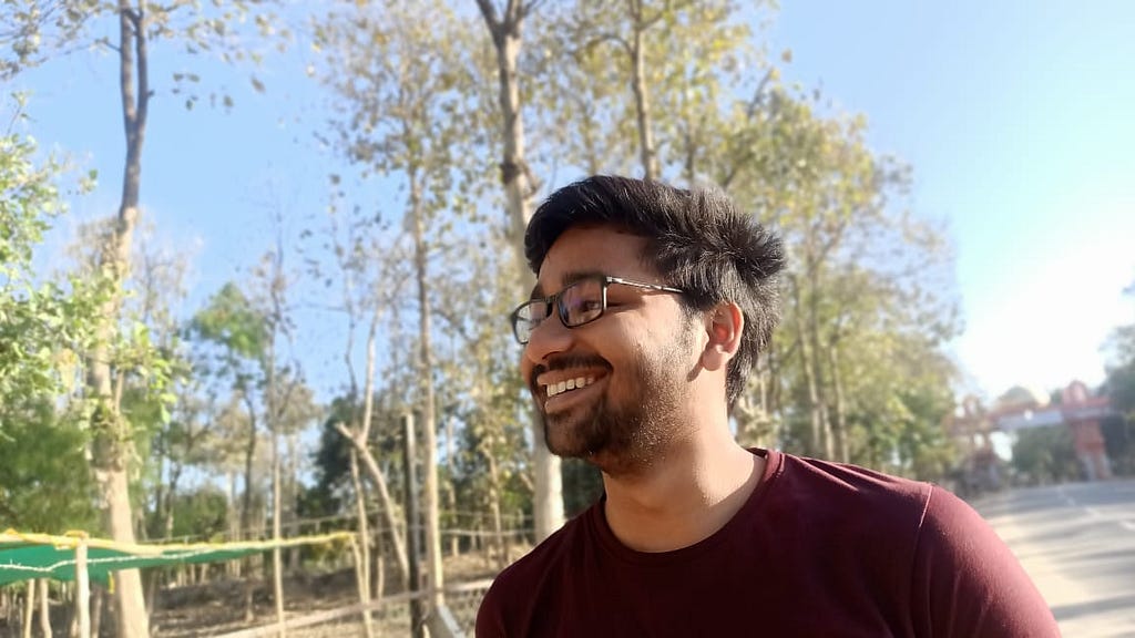 Photo of an Indian man, who is smiling and wearing a red shirt with black hair and spectacles. He is standing at a temple’s gate near a mild dense forest.
