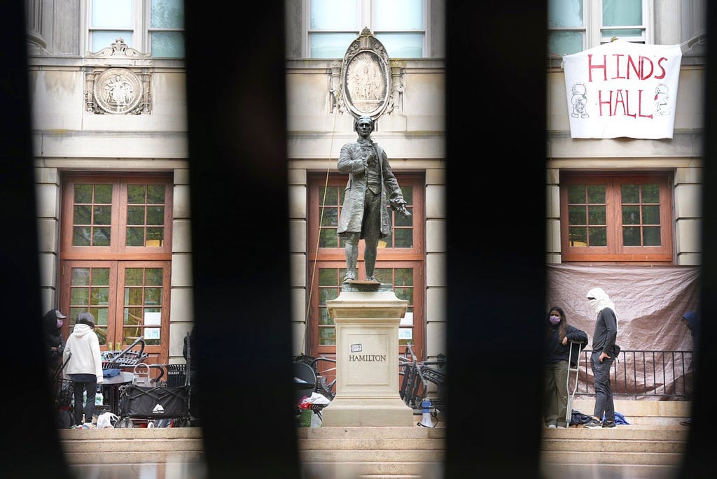 A statue of a person is viewed behind bars which obscure the camera’s view. A makeshift white sign with red lettering drapes from the building reading “HINDS HALL.”