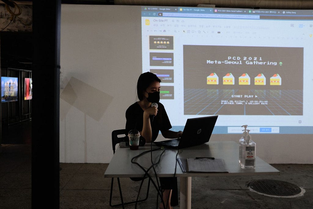 A woman sits at a table with a laptop. Behind her is a projection with a slide that says, “PCD 2021 Meta-Seoul Gathering” with illustrations of slices of cake.