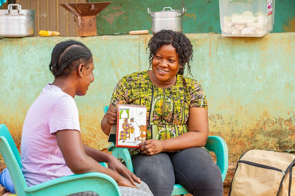 Judith sitting and talking to another sitting woman. Judith is holding a contraceptives information flip book and showing it to the other woman.