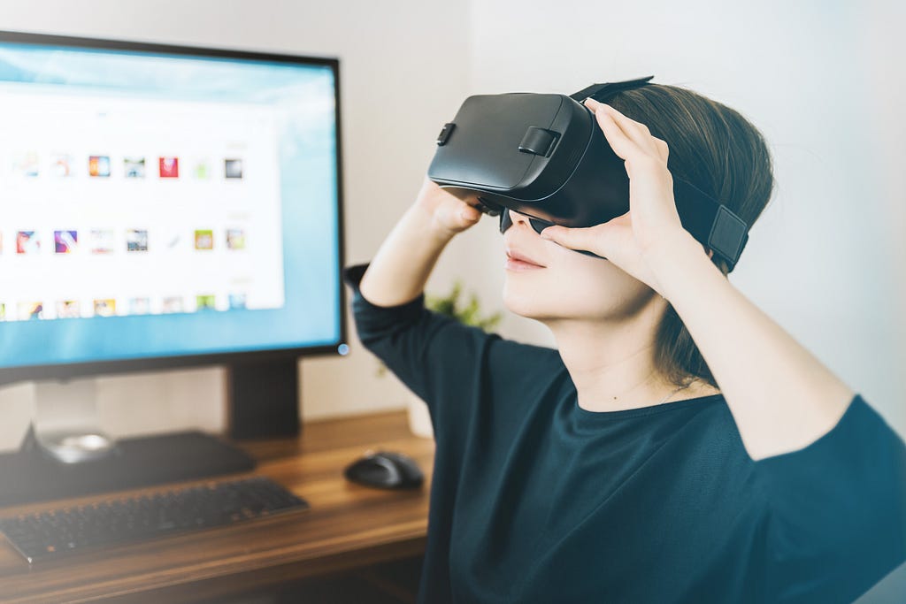 Young adult in black shirt sits in front of a computer monitor wearing virtual reality headset.