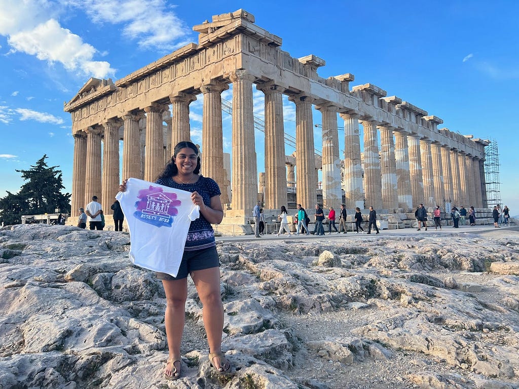 An image of a girl (the author of this post) standing in front of the Parthenon holding an ASSETS ’22 T-shirt.