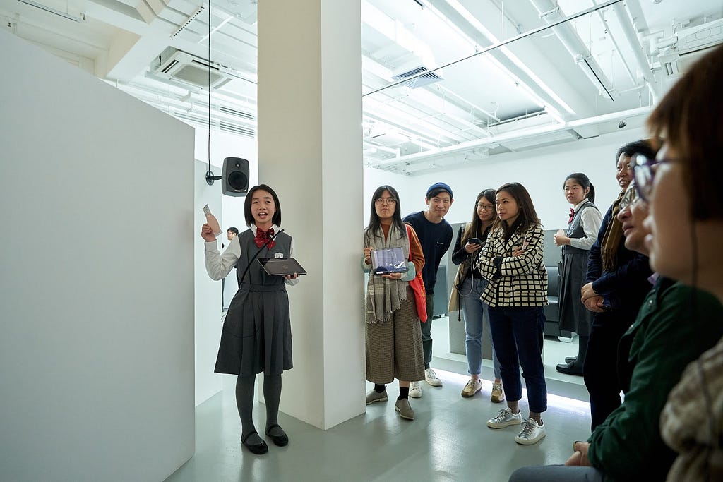 一個年幼學生站在展廳中，向圍觀的一群人說話，伸手指向身後的白色牆壁。