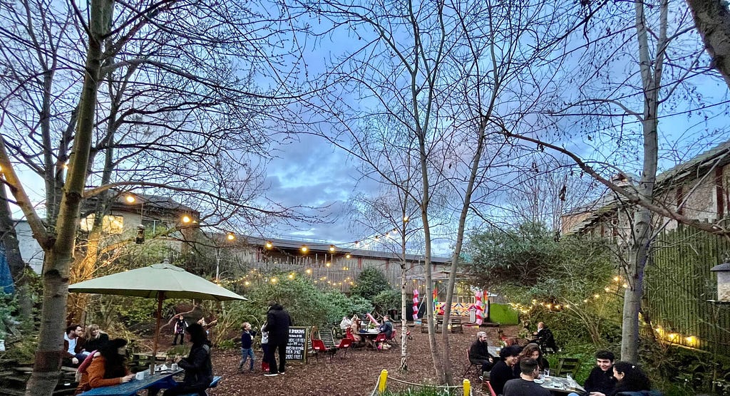 A natural city garden with trees, fairy lights and people sitting at tables