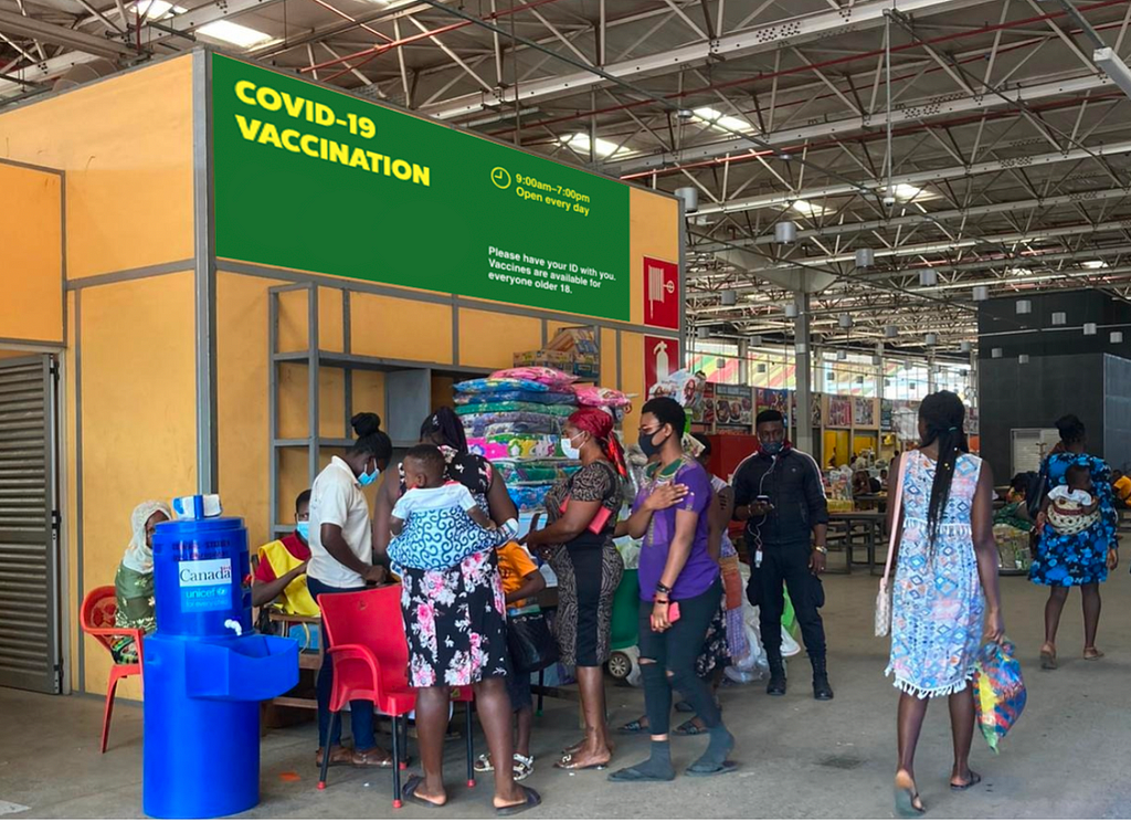 Large green banner above pop-up vaccination site in the marketplace that reaads “COVID-19 VACCINATION” in yellow text. The sign features the pop-up’s hours of operation, and states “Please have your ID with you. Vaccines are available to everyone older 18.”