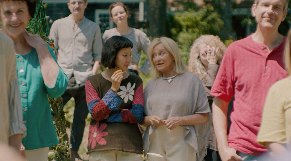 Maggie is holding a cherry tomato. Her head is turned towards an older resident who reminds her of the communal eating rules. There are 7 other artists standing in frame and listening to the orientation. All except Maggie are white.