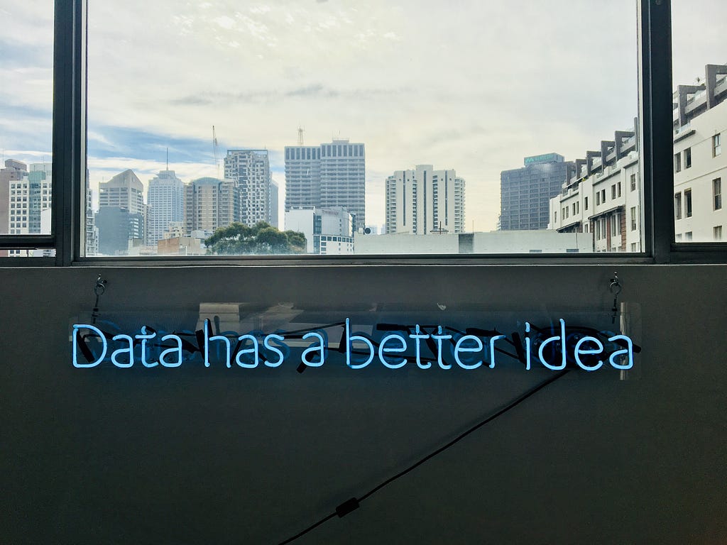 Neon Blue lettering. Phrase is ‘Data has a better idea’. It is below a window which has a view of a city skyline behind it.